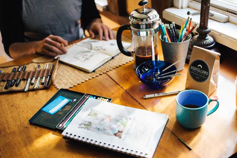 Two sketchbooks on a table next to a coffee mug, drawing utensils, with a person about to draw on one of the sketchbooks.